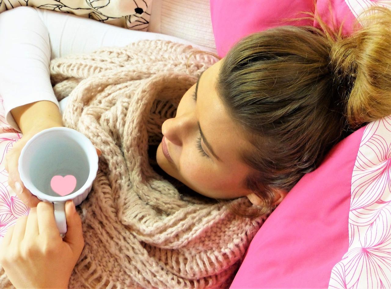 Kranke junge Frau im Bett mit einer Tasse Tee und einem Schal um den Hals.