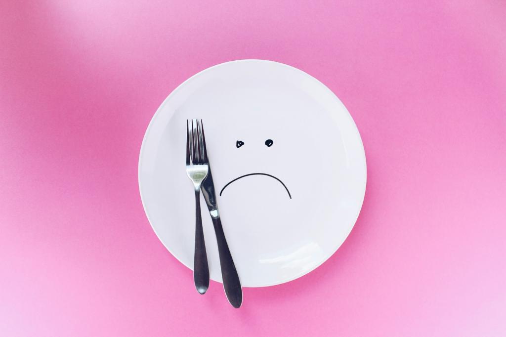 Leerer Teller mit Besteck und traurigem Smiley vor rosa Hintergrund.